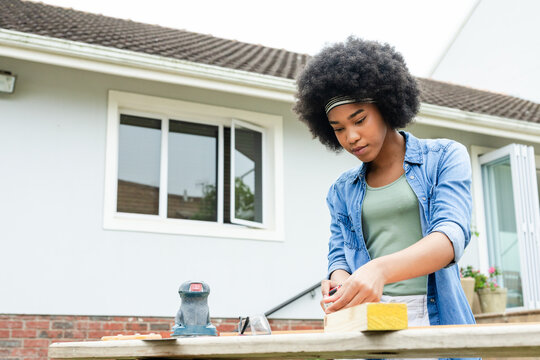 African American woman in denim shirt working outside suburban house marking wood plank with sander