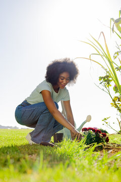 Woman crouching, planting red flowers in patch on lawn, wearing green tee, watering can nearby