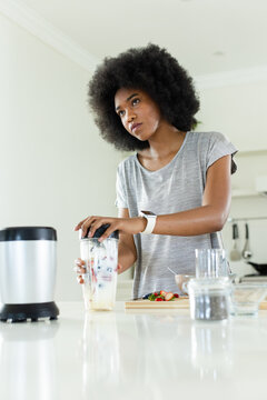 African woman standing at kitchen counter holding clear cup, securing lid beside blender base