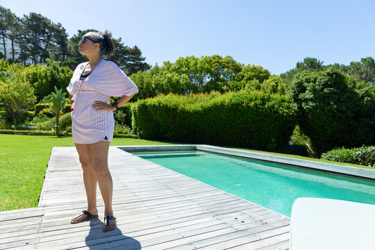 Sunlight falling across deck, woman standing hands on hips, looking up left, surveying pool area