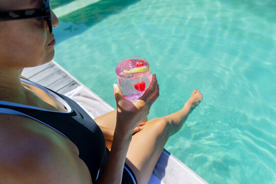 Sun urging woman at pool sipping pink drink, dipping leg, animated tags circling glass boosting ads