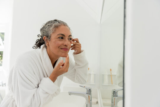 Senior African American woman leaning at bathroom sink mirror, applying cream near eye in bathrobe
