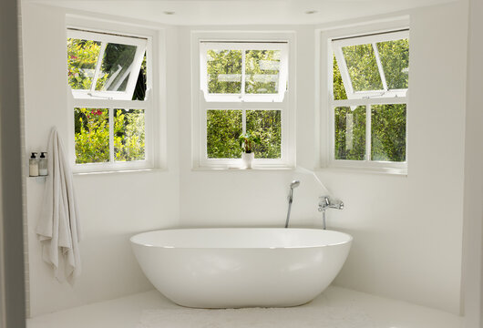 Freestanding oval bathtub sitting beneath three casement windows, showing chrome faucet and towels