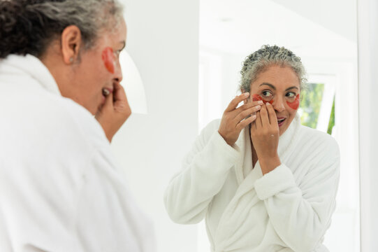 Senior African American woman applying red under-eye patches in white bathrobe at bathroom mirror