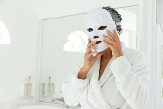 Woman starting LED therapy by lifting white facial mask and adjusting strap for skin care