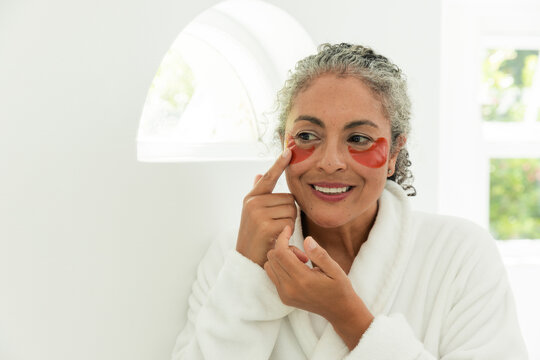 African woman applying red gel eye patches, wearing white soft robe in bathroom near window plants
