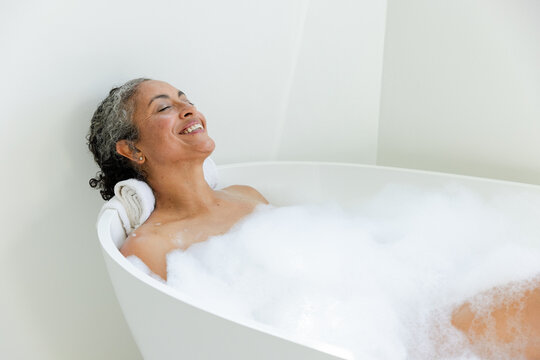 Senior African American woman reclining in tub in bathroom, foam, towel headrest and stud visible