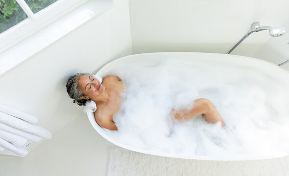 African American woman reclining in bathroom tub, resting head on rolled towel with bubble foam