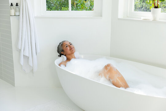 Senior African American woman reclining in bright bathroom in freestanding bathtub with foam