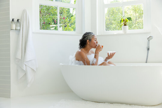 Vertical video: Sipping senior woman soaking in oval tub, light through windows with cup and saucer