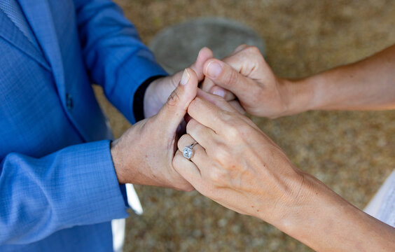 Couple holding hands with blue suit, white dress showing diamond ring on left finger over gravel