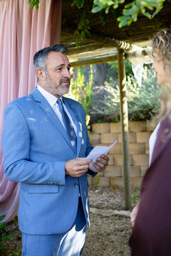 Middle-aged couple reading vows under pergola with pink drape, man in light-blue suit holding paper
