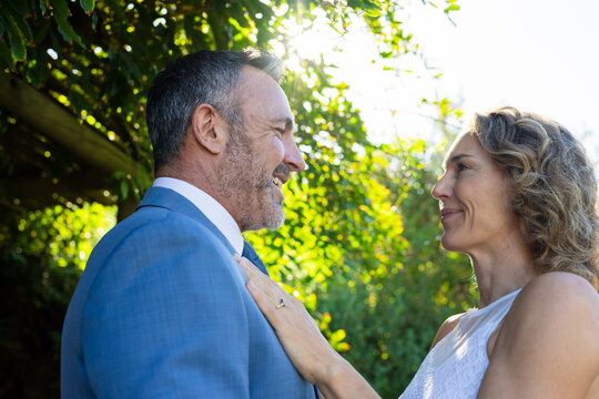 Couple in formal wear standing close in garden under pergola, showing engagement ring