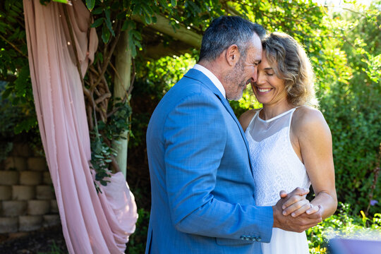 Couple standing under arbor with pink drape in garden pressing foreheads in blue suit, white dress