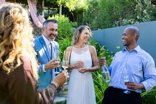 Diverse friends woman in white lace dress man in blue suit raising champagne flutes in garden