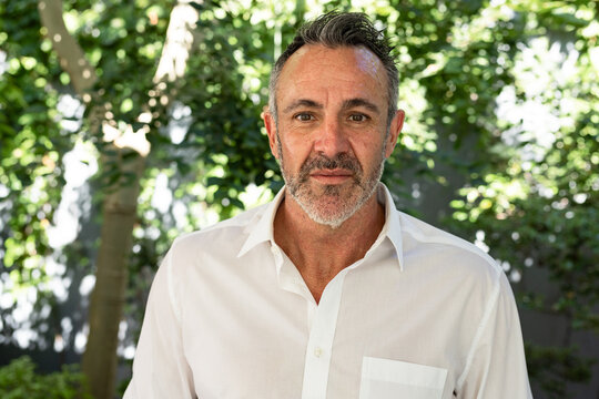 Senior man standing in backyard by tree trunk wearing white button-down shirt with dappled sunlight