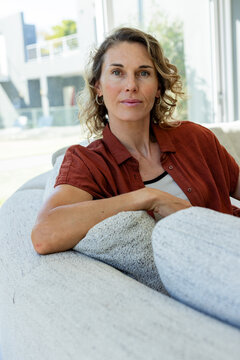Woman sitting on light-gray sofa in living room by large windows, wearing rust shirt