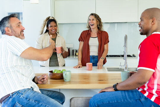 Diverse friends chatting and laughing at kitchen island holding mugs near bowl of green apples