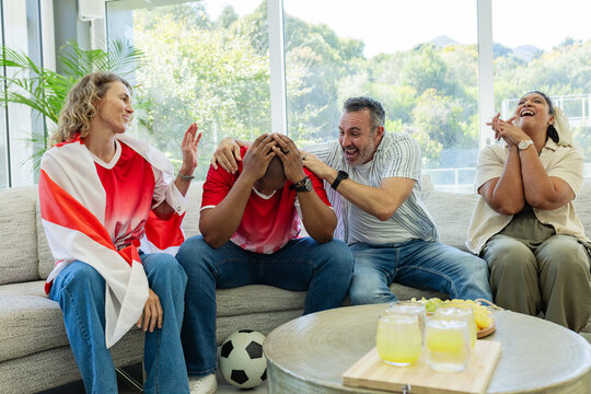 Diverse friends reacting to soccer match in living room on sofa with soccer ball, red jerseys