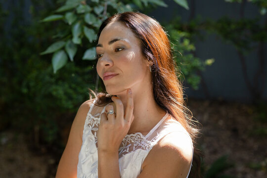 Adult woman posing in backyard garden touching chin wearing white lace sleeveless top, silver ring