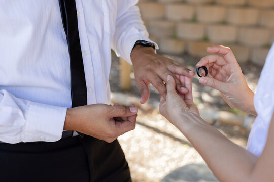 Couple exchanging small dark ring in courtyard, white shirt, black tie, dark wristwatch