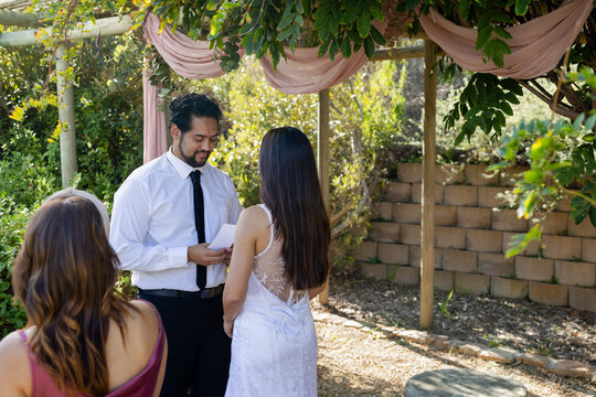 Bride in white lace dress and officiant in black tie standing under pergola holding folded paper