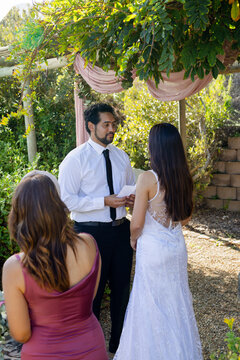 Couple exchanging vows under pergola in garden, man holding paper, woman wearing lace gown