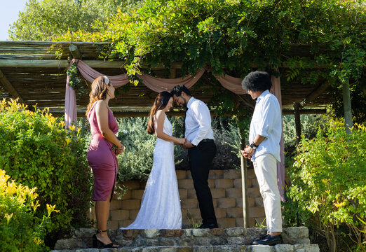 Wedding couple holding hands, heads touching under pergola on stone platform, attendants with post