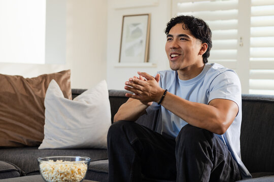 Man sitting on gray sofa in living room clapping, wearing blue tee and bracelet, popcorn nearby