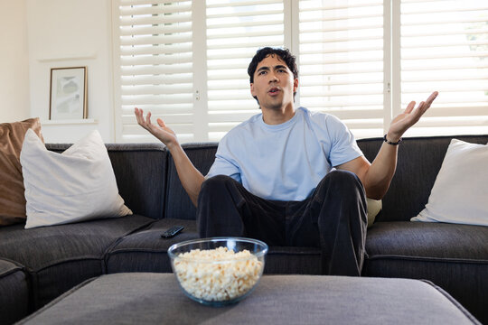 Asian man sitting on sofa in living room raising arms, watching TV with popcorn and remote