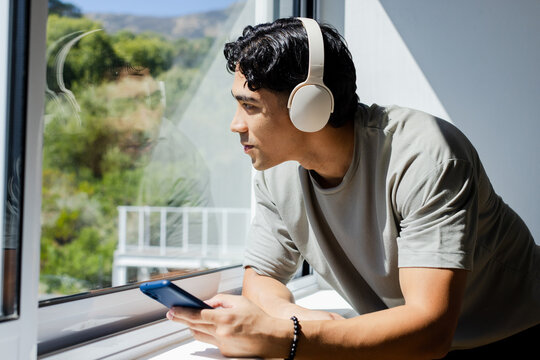 Asian man leaning on windowsill and looking outside, bright room with white headset and blue phone