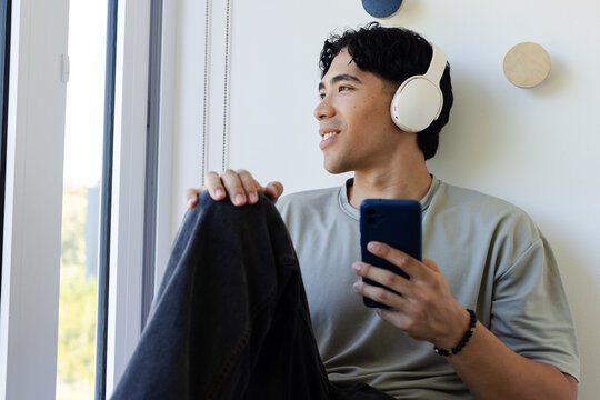 Adult Asian man sitting on cushioned window seat at window, wearing headphones, holding blue phone