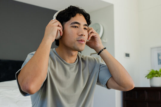 Asian man adjusting white over-ear headphones while wearing grey tee and black bracelet in bedroom