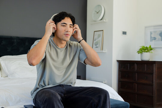 Asian adult man sitting on bed adjusting over-ear headphones while checking phone near headboard