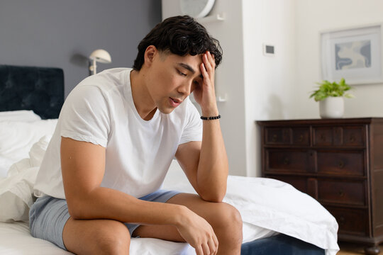 Asian man sitting on bed in bedroom, leaning forward, pressing hand to forehead, wearing white tee