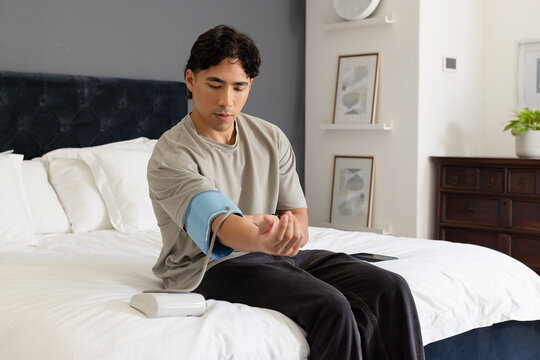 Asian adult man sitting on bed in bedroom, connecting monitor and wrapping blood pressure cuff