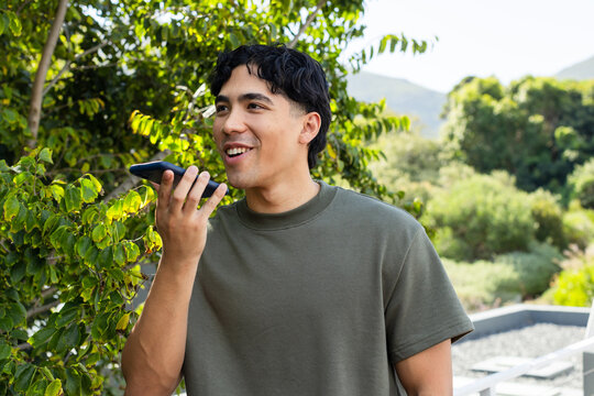Adult male standing on terrace wearing olive-green T-shirt, holding phone near mouth by shrubs