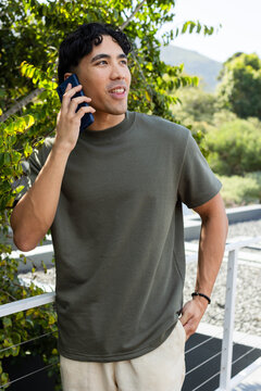 Asian man standing on balcony holding smartphone to ear by metal railing, wearing olive tee