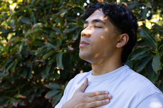 Male standing on right amidst sunlit shrubs with hand resting on chest and crewneck shirt