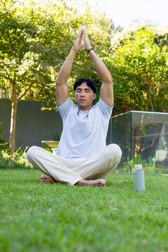Sitting man wearing light tee, meditating arms-up, relaxing with sunlight filtering, bottle right