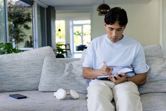 Adult Asian man writing in notebook on sofa at home wearing blue tee, smartphone, copy space