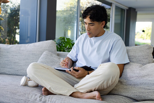 Male in twenties sitting on sofa in living room writing in notebook with pen, phone nearby