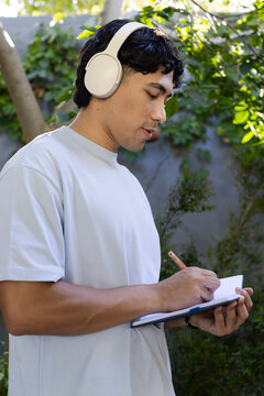 Male student wearing over-ear headphones, hearing lecture and writing notes in sunny backyard study