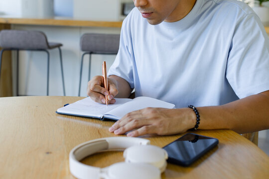 Asian man sitting at kitchen table writing in open notebook with pen, smartphone, headphones