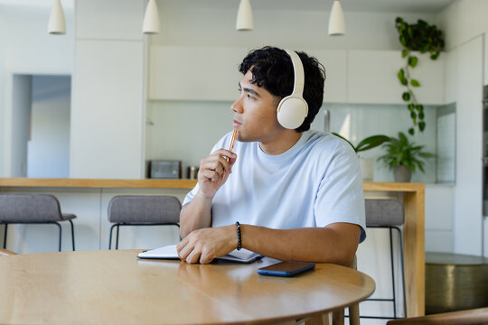 Asian adult man wearing headphones, holding pen at open notebook and phone at round-wooden table