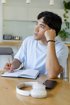 Asian man sitting at wooden table in kitchen, writing notebook with pen, headphones, light-blue tee