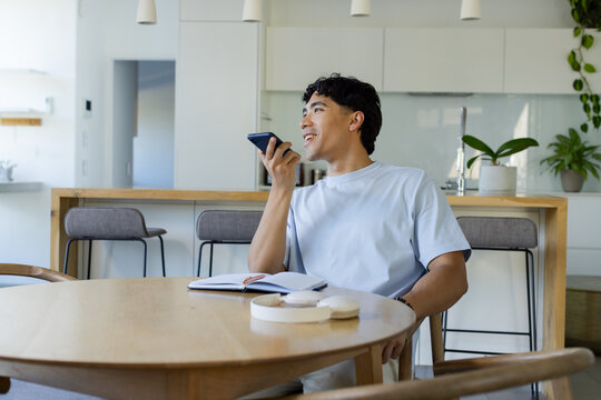 Asian man sitting at dining table in kitchen, speaking into phone with notebook and earbud case