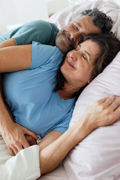 Couple lying in bed embracing, resting in bedroom with pale pillows, duvet, short-sleeve T-shirts