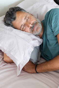 Mature man sleeping on side in bed with pale sheets, pillow, wearing teal-green T-shirt
