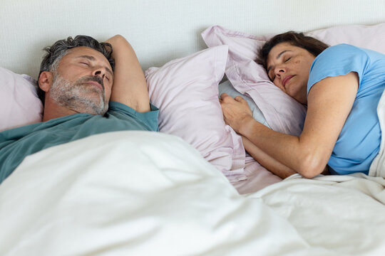 Senior couple lying in bed in bedroom under white duvet, pale pink pillows, wearing short sleeves